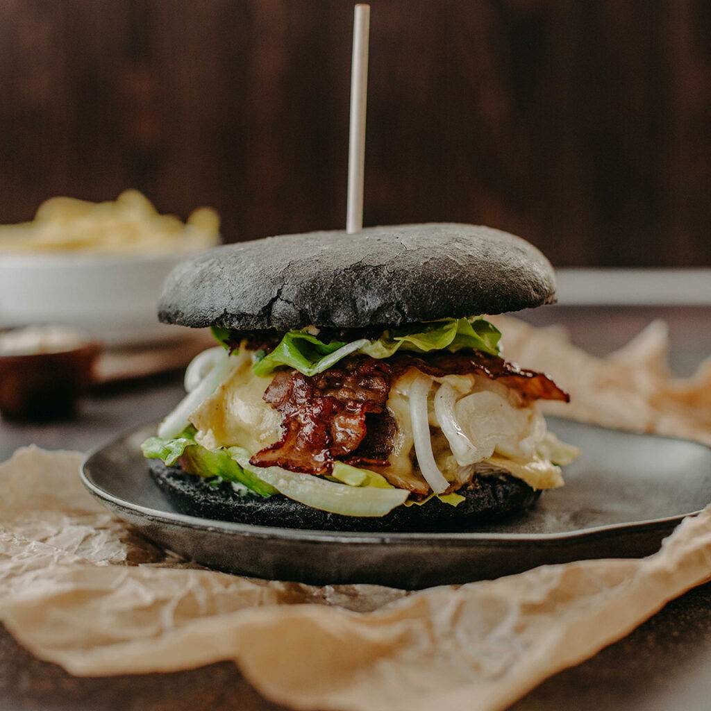 Ein Gourmet-Burger mit schwarzem Brötchen, belegt mit geschmolzenem Käse, Speckstreifen, Salat und Zwiebeln, serviert auf einem dunklen Teller in diesem exquisiten Restaurant. Der Burger ist mit einem Spieß zusammengehalten, und vor dem unscharfen Hintergrund ist eine Schüssel Pommes Frites zu sehen.