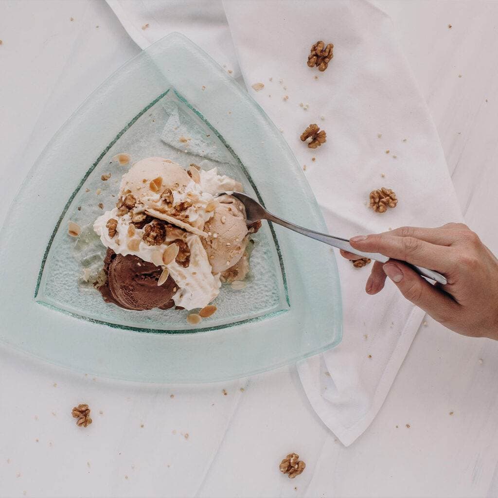 Eine Hand hält einen Löffel und greift gerade in einen dreieckigen Glasteller mit Eiscreme in einem Restaurant. Das Eis besteht aus mehreren Kugeln, die mit Schlagsahne, zerstoßenen Nüssen und Walnüssen garniert sind. Auf der weißen Oberfläche sind verstreute Nüsse und ein weißes Tuch zu sehen.