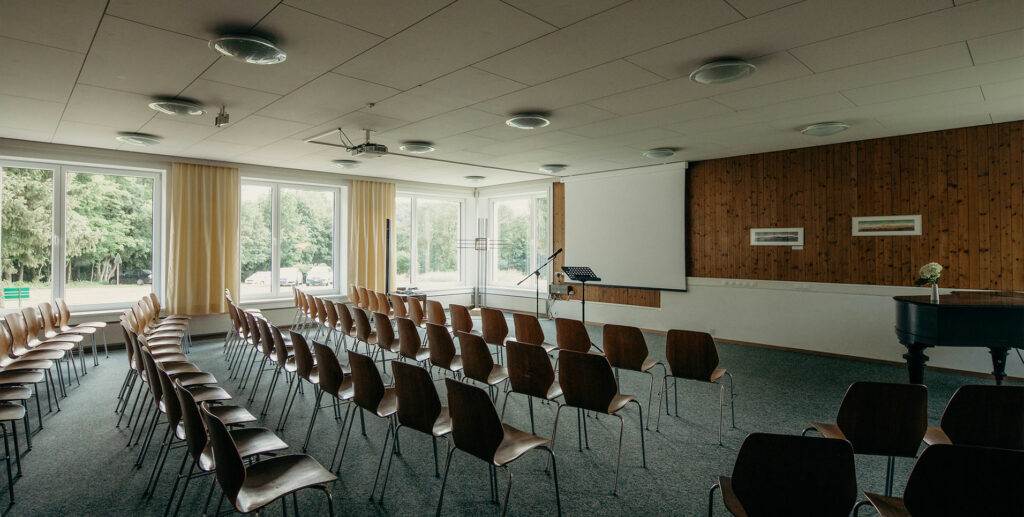 Ein leerer Konferenzraum mit Reihen brauner Stühle vor einer großen Projektionsleinwand, perfekt für ein Seminar. Der Raum hat große Fenster, die Tageslicht hereinlassen, einen Flügel in der Ecke und eine Holzwand mit zwei gerahmten Bildern. Der Raum wirkt sauber und ordentlich.