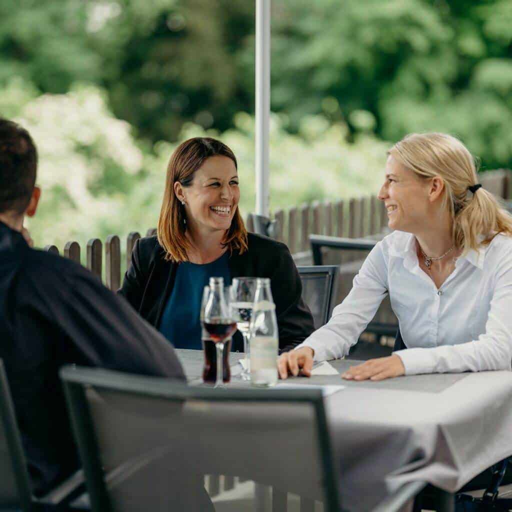 Drei Personen sitzen an einem Tisch im Freien, unterhalten sich und lächeln. Der Hintergrund ist üppig und grün und lässt eine Gartenkulisse vermuten, die an eine ruhige Galerie erinnert. Zwei Frauen, eine mit dunklem Haar und eine mit blondem Haar, stehen sich gegenüber, während ein Mann mit dunklem Haar in der Nähe sitzt.