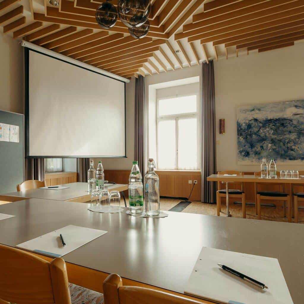 Ein Konferenzraum mit einer großen Projektionsleinwand, einem Tisch mit Stühlen, Wasserflaschen und Gläsern. Der Raum hat eine moderne Holzdeckengestaltung, die an eine Kunstgalerie erinnert, Kunstwerke an den Wänden wie bei einer Ausstellung in einer Galerie und ein großes Fenster, durch das natürliches Licht hereinfällt. Notizblöcke und Stifte liegen auf dem Tisch.