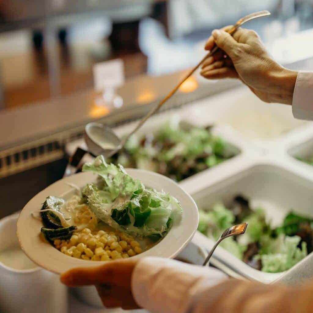 In einem Kunstgalerie-Café hält eine Person einen weißen Teller mit Salat, Mais und anderem Gemüse vom Salatbuffet. Mit einer Schöpfkelle gießt sie cremiges Dressing über den bunten Salat. Im Hintergrund sind verschiedene Behälter mit frischen Zutaten zu sehen, die an eine essbare Ausstellung erinnern.