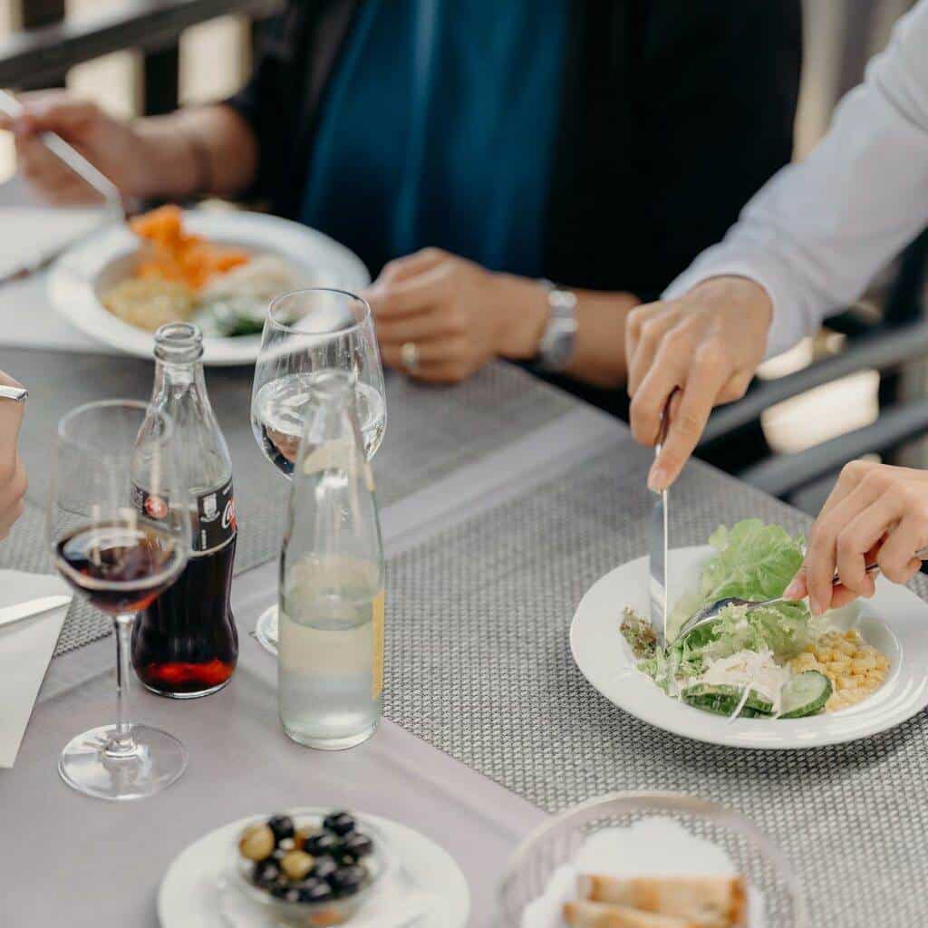 Drei Personen essen an einem Tisch mit grauen Tischsets, der an eine Ausstellung erinnert. Sie genießen verschiedene Gerichte, darunter einen Salat und einen Teller Pasta, dazu Gläser Rotwein, Limonade und Mineralwasser. Auf dem Tisch stehen außerdem Brot und eine Schale Oliven. Hände und Geschirr sind zu sehen.