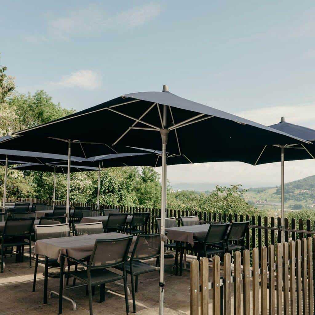 Eine Restaurantterrasse im Freien mit Tischen und Stühlen unter großen marineblauen Sonnenschirmen. Der Bereich ist von einem Holzzaun und üppigem Grün umgeben, was eine ruhige, galerieartige Atmosphäre schafft. Im Hintergrund ist eine malerische Aussicht auf eine ferne Landschaft mit Hügeln und Bäumen unter einem blauen Himmel mit wenigen Wolken zu sehen.