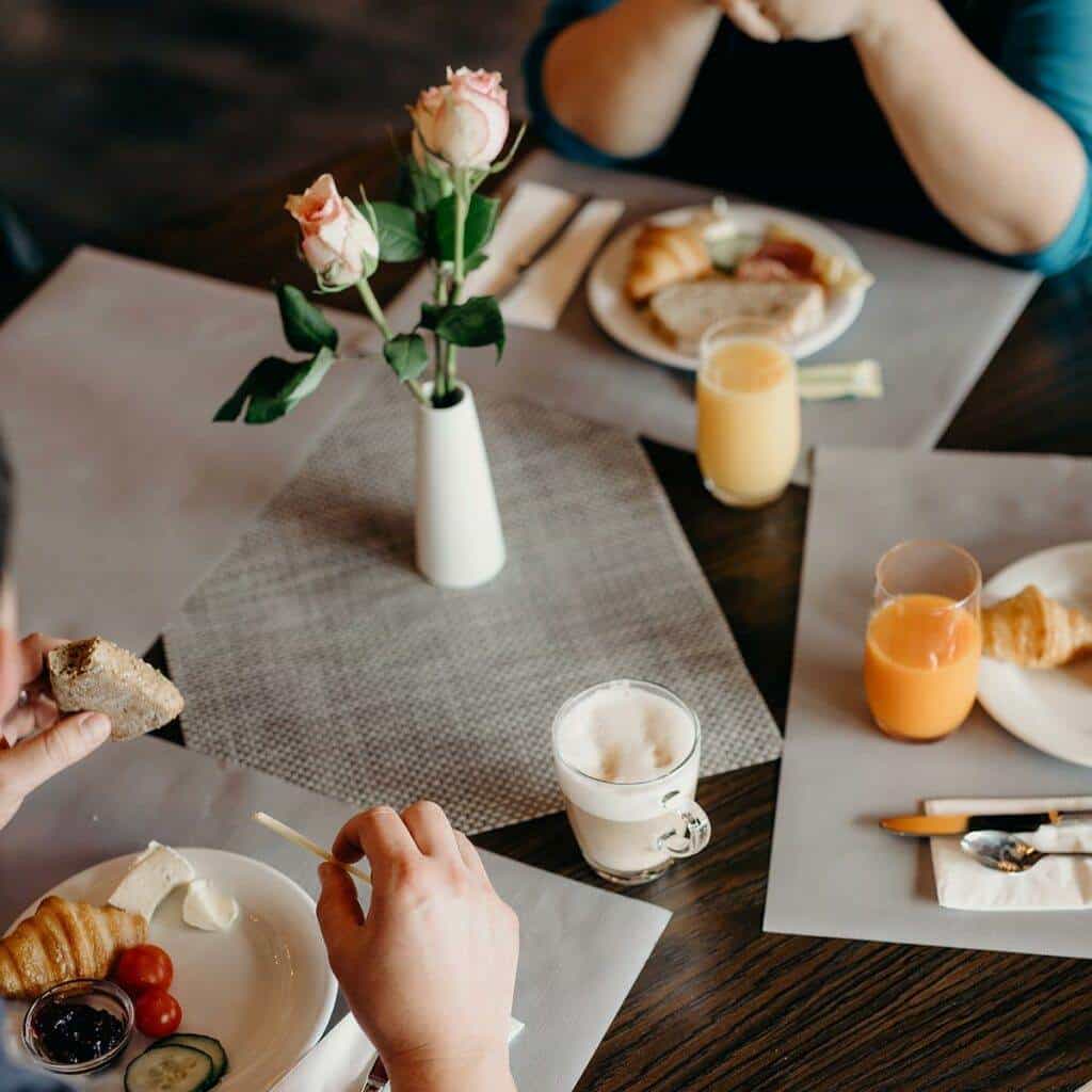 Zwei Personen sitzen an einem Tisch und genießen ihr Frühstück, das Croissants, Brot, Käse, Kirschtomaten und Gurken umfasst. Sie haben Gläser mit Orangensaft und eine Tasse schaumigen Kaffee. In der Mitte des Tisches steht eine Vase mit Rosen. Die Stimmung ist gemütlich und ungezwungen, als wären sie im Café einer Kunstgalerie.