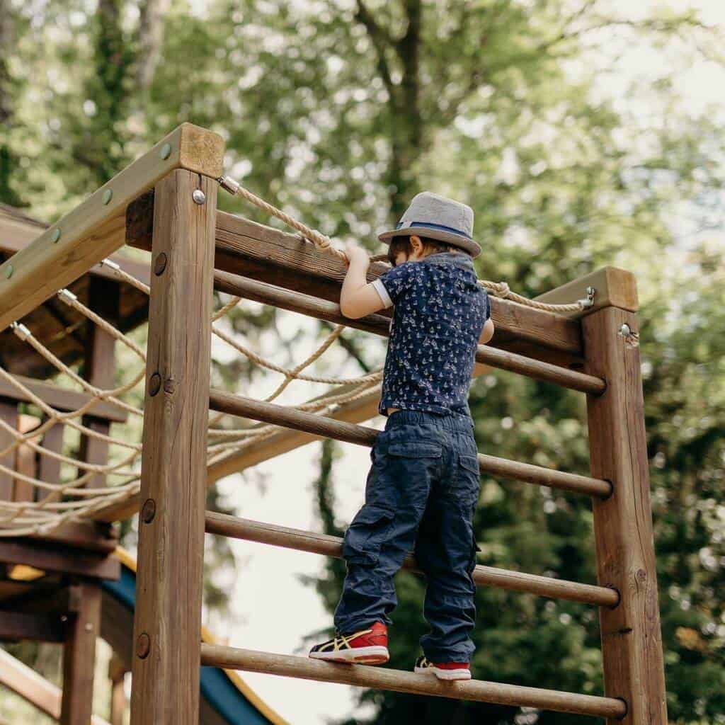 Ein Kind mit Hut, marineblauem Hemd und blauer Hose klettert auf einem von Grün und Bäumen umgebenen Spielplatz im Freien auf ein hölzernes Klettergerüst mit Seil- und Leiterelementen. Es ist wie eine lebendige Galerie spielerischer Abenteuer inmitten der Natur.