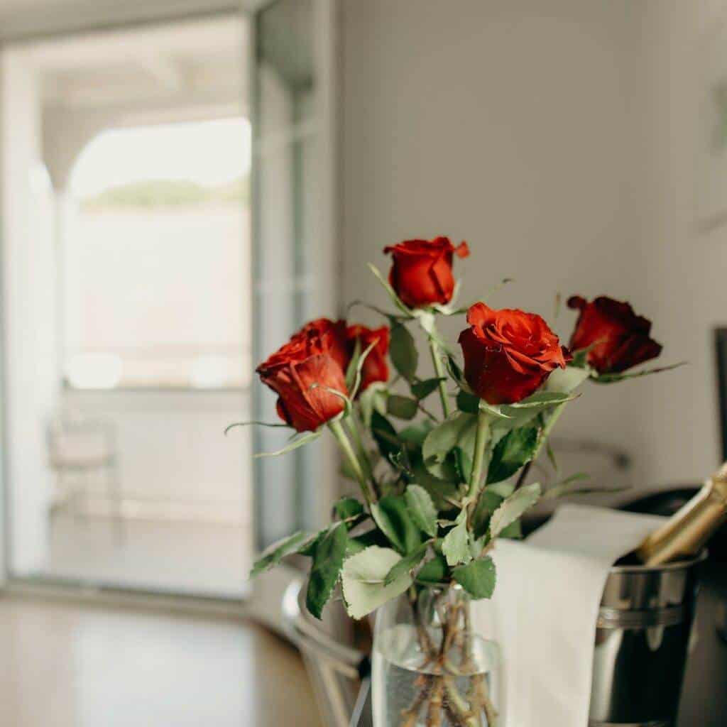 Ein Strauß roter Rosen in einer Glasvase steht auf einem Tisch in einem gut beleuchteten Raum. Eine Flasche Champagner in einem Eiskübel steht neben der Vase. Im Hintergrund öffnet sich eine Glasschiebetür zu einem Balkon mit einem Metallstuhl, der an eine elegante Szene aus einer Kunstgalerie erinnert.