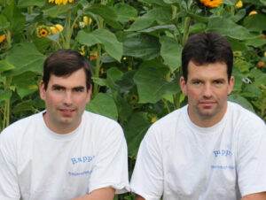 Deux hommes en chemise blanche avec un texte bleu sur le côté gauche sont assis devant un fond de feuillage vert et de fleurs jaunes symbolisant la durabilité. Ils ont tous deux les cheveux courts et foncés et regardent directement la caméra.