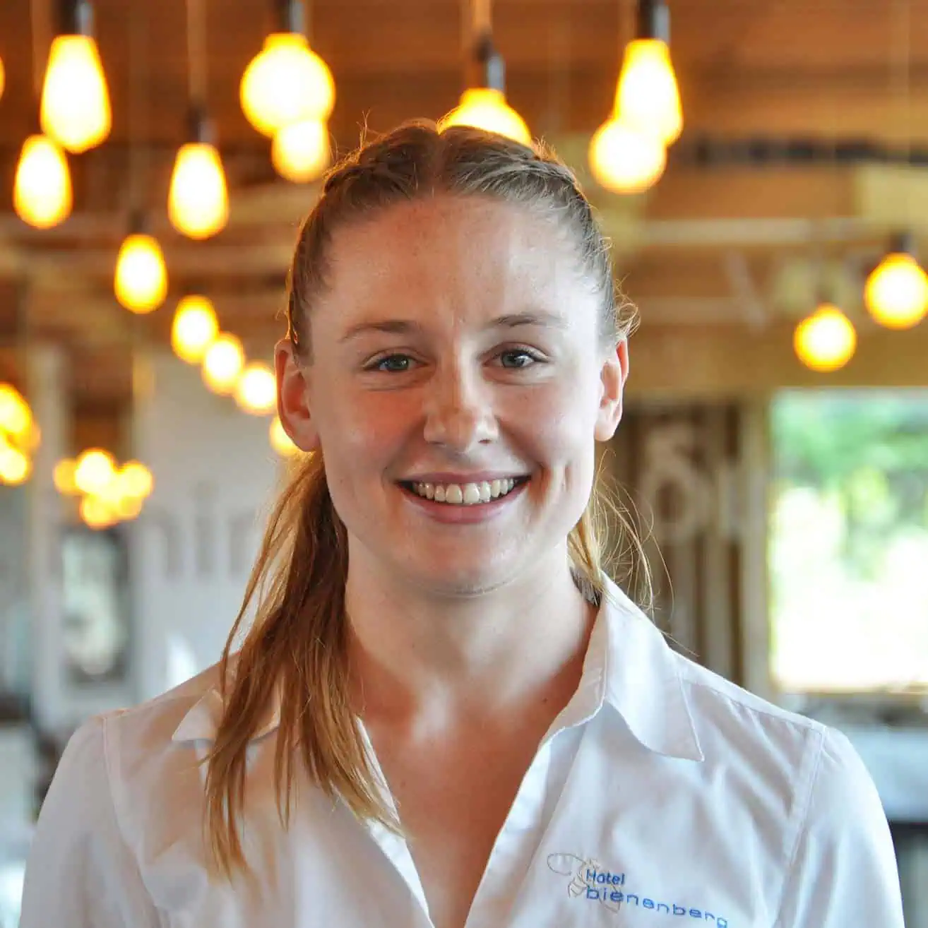 A smiling woman with long hair tied back stands in a warmly lit interior. She is wearing a white button-down shirt adorned with the "Hotel Bienenberg" logo. Several hanging lamps can be seen in the background, creating an inviting atmosphere that reflects our "About us" hospitality philosophy.