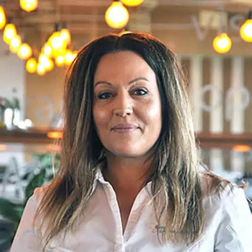 A person with long, dark hair stands in an indoor area wearing a white shirt. In the background, hanging lights illuminate the blurred elements of a restaurant or café, creating a cozy scene reminiscent of an inviting "About Us" page.