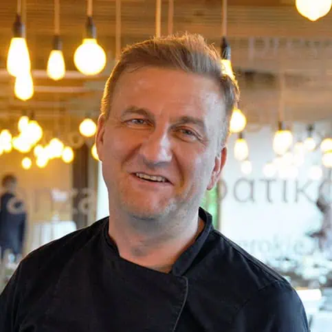 A smiling man with short, light-colored hair and a dark shirt stands in a room with warm, hanging light bulbs in the background. The setting looks cozy and well-lit, creating a perfect atmosphere for an "About Us" introduction.