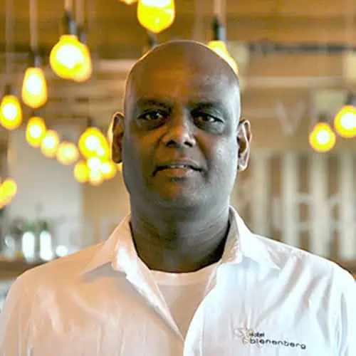 A person in a white shirt is standing in an indoor area, with several light bulbs hanging in the background, which are lit up. The person is looking into the camera and appears to be in a well-lit area. They embody the clarity and concentration that we want to achieve in "About us".