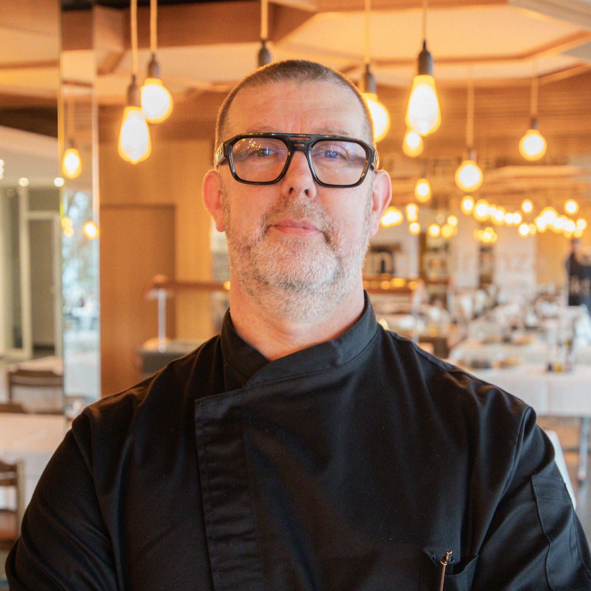 Ein schwarz gekleideter Koch mit Brille steht in einem warm beleuchteten Restaurant mit hängenden Glühbirnen. Das Ambiente ist elegant, mit weißen Tischdecken und Holzakzenten.